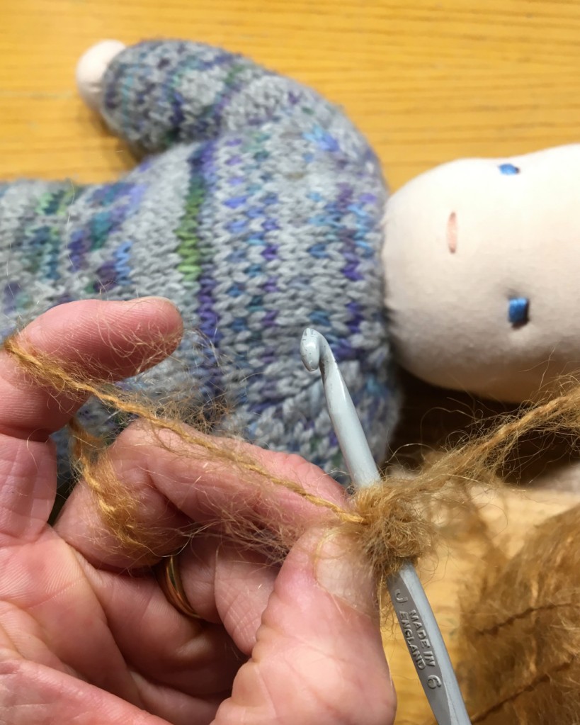 Image show a hand holding a crochet hook and fluffy light-brown yarn in the foreground. In the background there is a handmade doll with a knitted body, blue eyes and pink mouth but no hair yet.