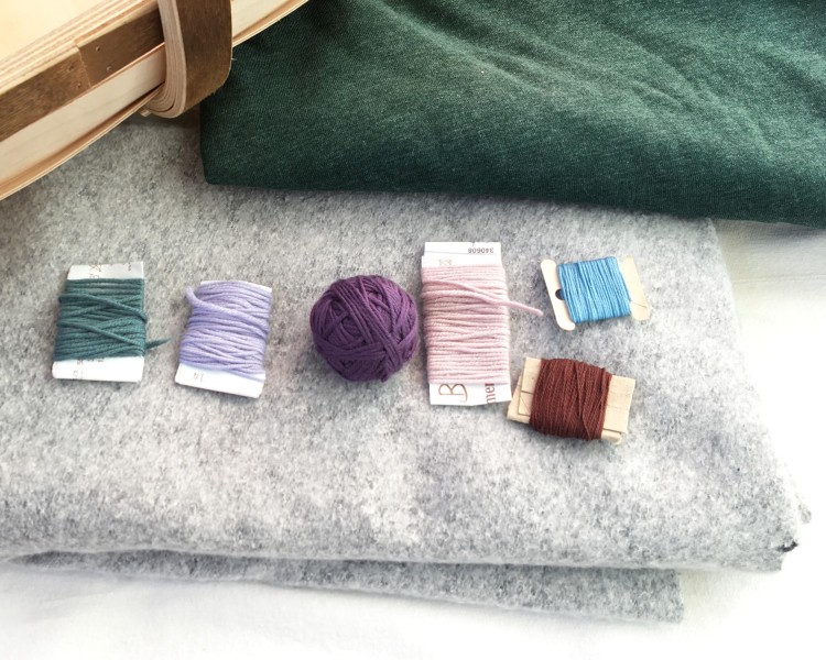 Image shows two pieces of folded jersey fabric in grey and green. On top of the grey fabric lie three bobbins of wool in green, blue and pink; a small ball of purple wool and two bobbins of embroidery thread in blue and brown.