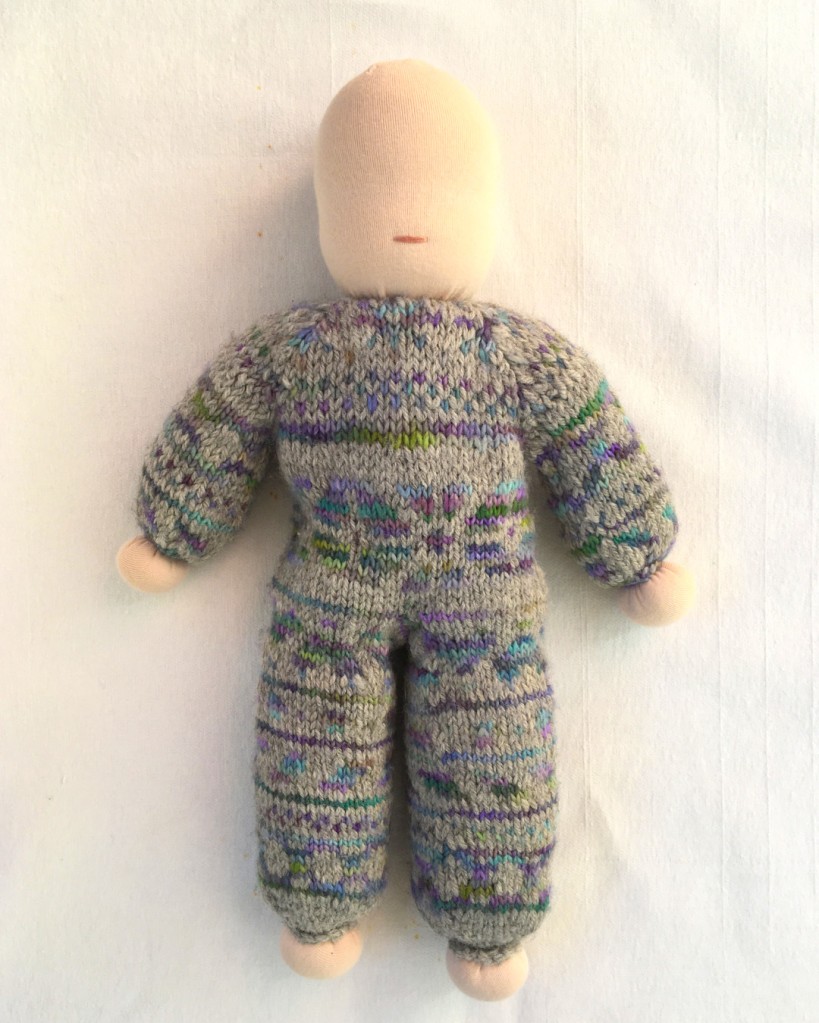 Image shows a handmade doll with knitted body in fair-isle design using grey, blue and green wool. The doll has simple hands and feet but an unfinished face. There are no eyes yet, just a mouth, and no hair.